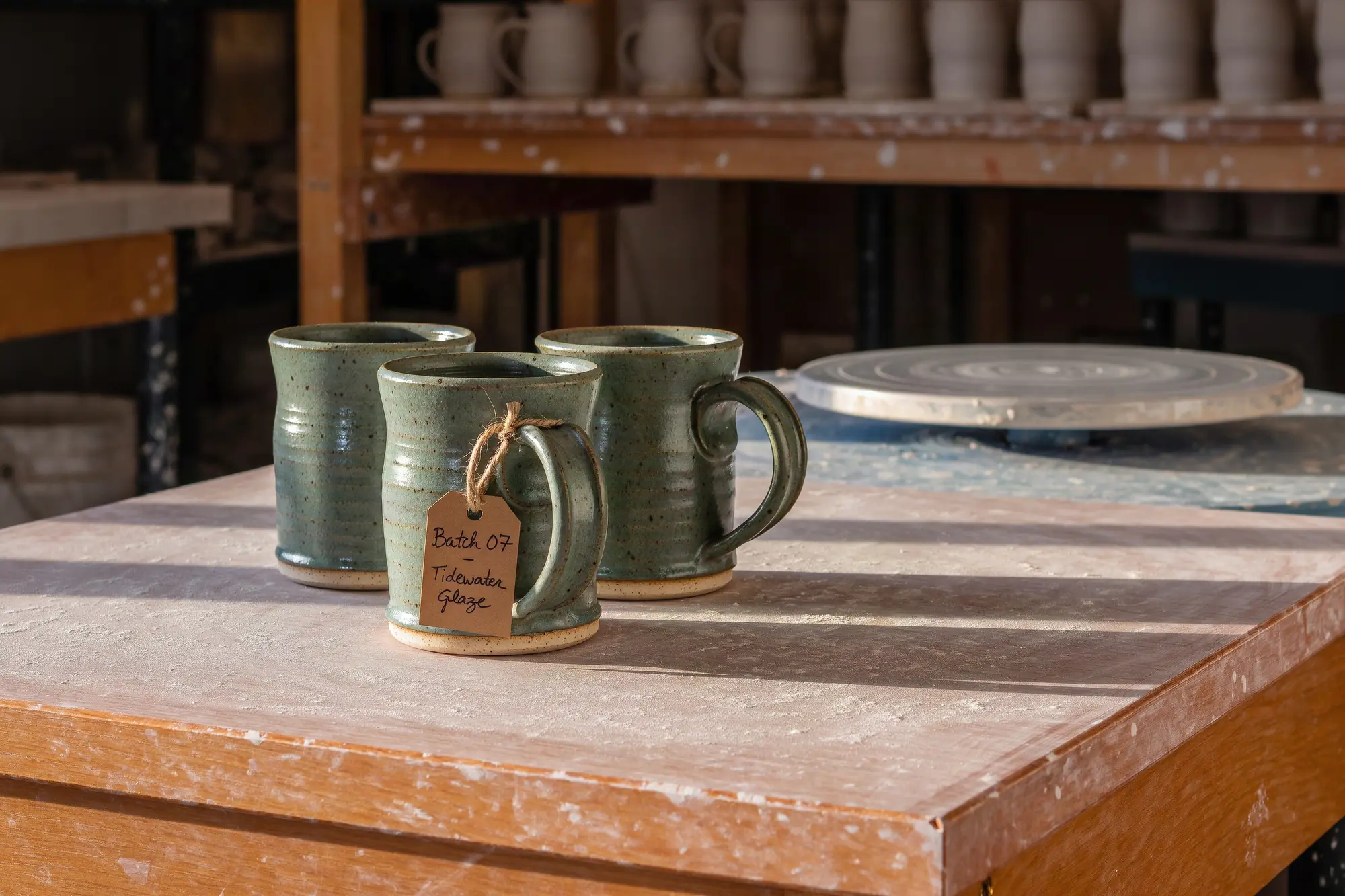 Krea 3 photo of three speckled sea-green mugs on a clay-dusted workbench in low sunlight, the front one tagged in handwriting Batch 07 - Tidewater Glaze